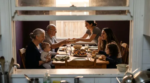 Three generations of a family around a shared table, the grandmother holding the baby in an everyday scene of alloparenting