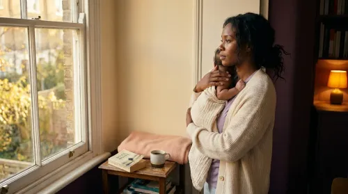 Black woman holding her infant at a window in late afternoon light, a quiet portrait among stories of motherhood