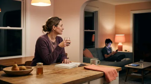 Mother reading at a wooden kitchen island in the evening with her teen on the living-room couch beyond, phone in hand