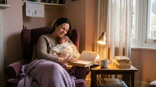 Mother in an armchair reading, her sleeping newborn against her shoulder — the unsleeping hours of novels about motherhood