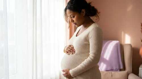 A woman cradling her pregnant belly in soft window light, both hands resting on a rounded bump in a cream knit top