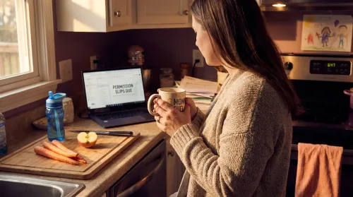 Working mother holding a "MOM FUEL" mug at her kitchen counter at 6:47pm, laptop open to "Permission Slips Due"