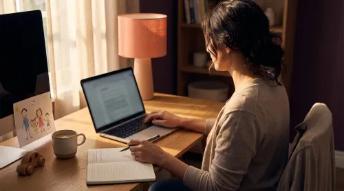 Mother at a home-office desk taking notes beside her laptop, a child's crayon drawing propped against the monitor
